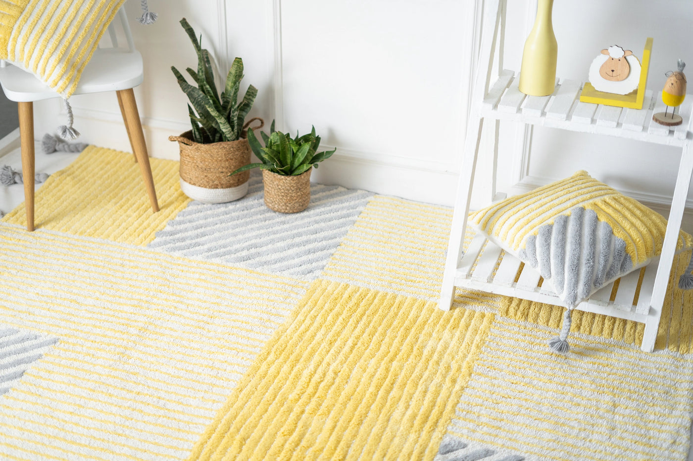 A cushion with yellow and grey stripes in various directions—vertical, horizontal, and diagonal—with tassels on all four corners, styled in a minimalist room featuring a butterfly rug.