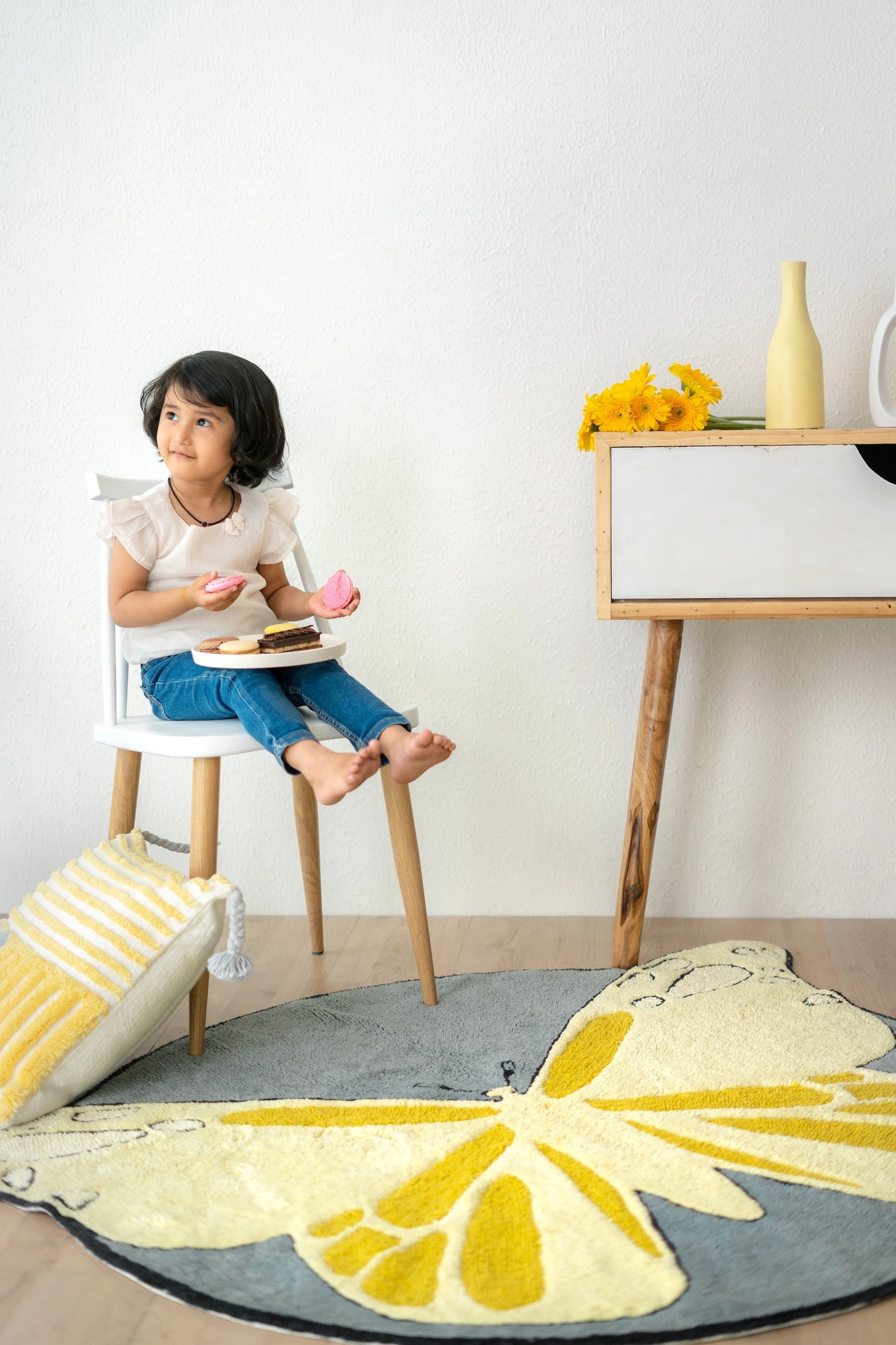 This cushion features yellow and grey stripes in vertical and horizontal patterns, with tassels on all four corners, placed in a minimalistic setting with a butterfly-patterned rug.