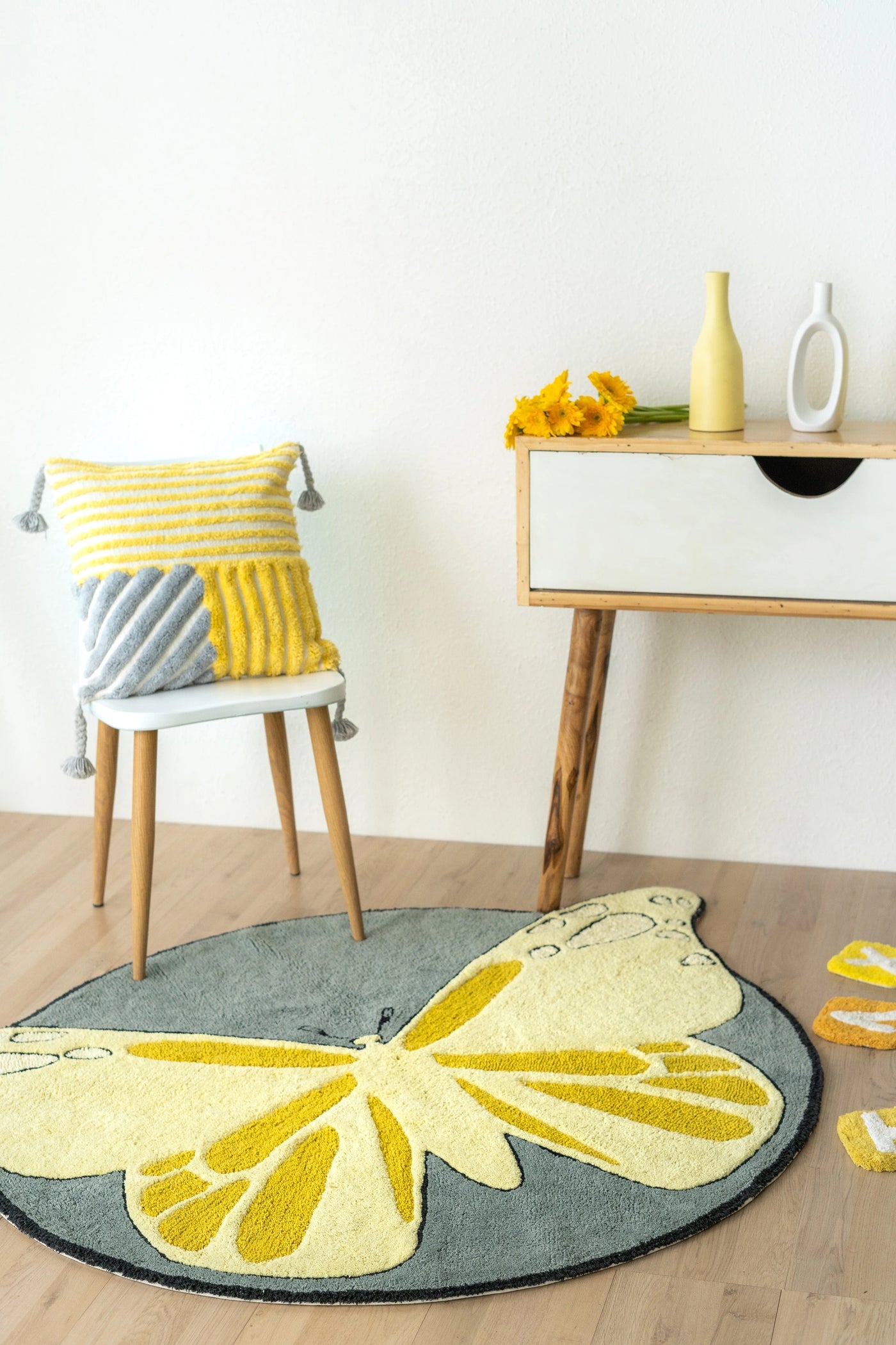 A striped yellow and grey cushion with vertical, horizontal, and diagonal yellow and grey stripes, featuring tassels on all four corners, with minimal interior and a butterfly-patterned rug.