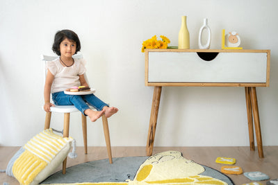 A yellow and grey striped cushion with vertical and horizontal stripes, accented with tassels on all corners, set against a minimal interior with a butterfly-patterned rug.